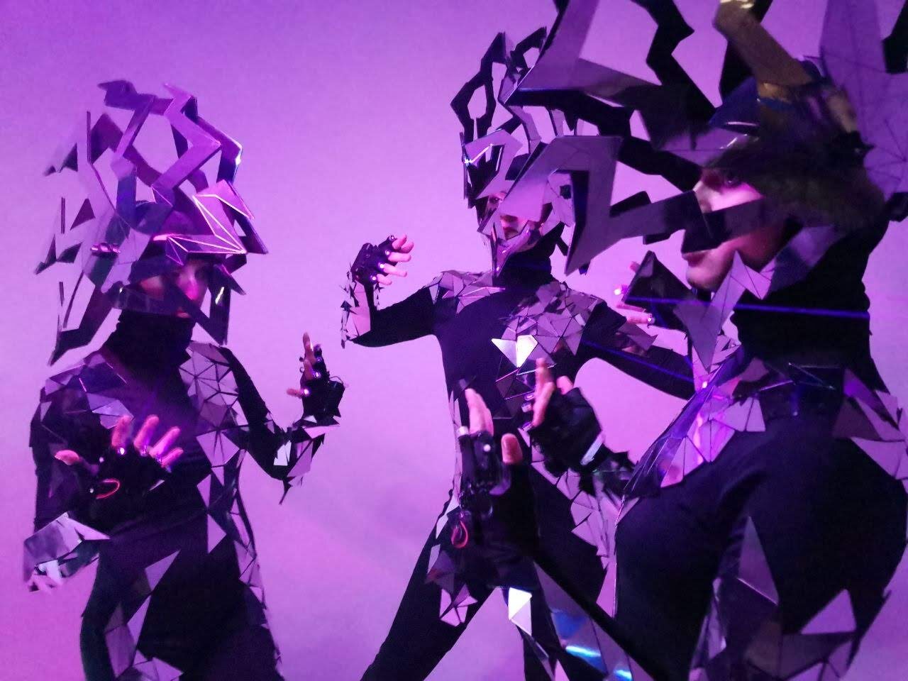 Three performers in mirror-mosaic costumes under purple stage light